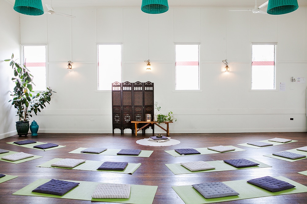 A bright, high-ceilinged wooden-floor studio set up for a yoga or meditation workshop, with rows of pale green yoga mats topped with folded meditation cushions arranged in a loose grid. At the front of the room, a dark wooden folding screen and a small low table with plants and candles form a focal point, flanked by tall windows letting abundant natural light in. Three teal woven pendant lights and ceiling fans hang from the high white ceiling, and a large potted plant stands in the left corner.
